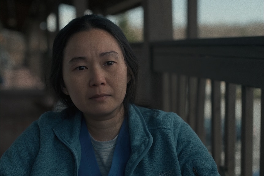 Middle-aged Asian woman with brown eyes and dark hair pulled back in a ponytail wears a blue jacket while sitting on a porch.