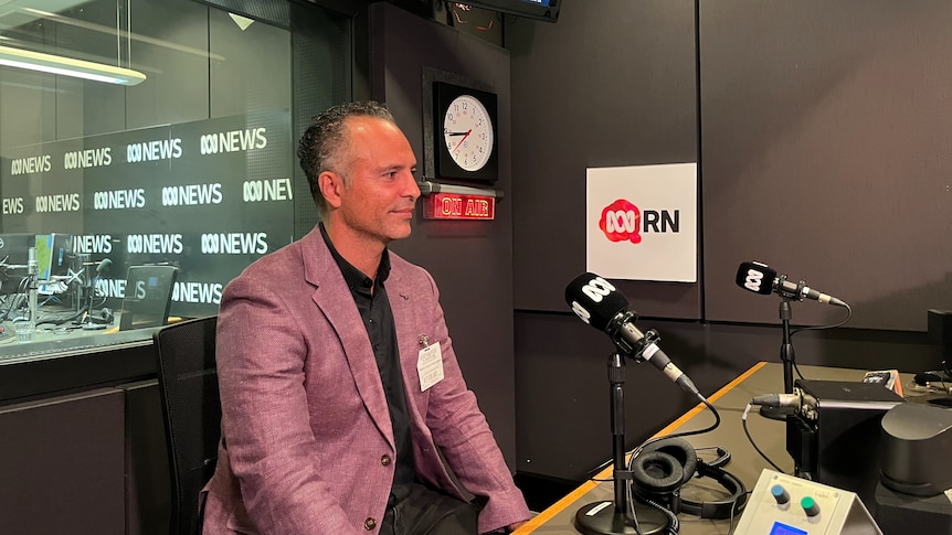 man in a pink linen blazer sits behind a microphone and looks forward, he is smiling 