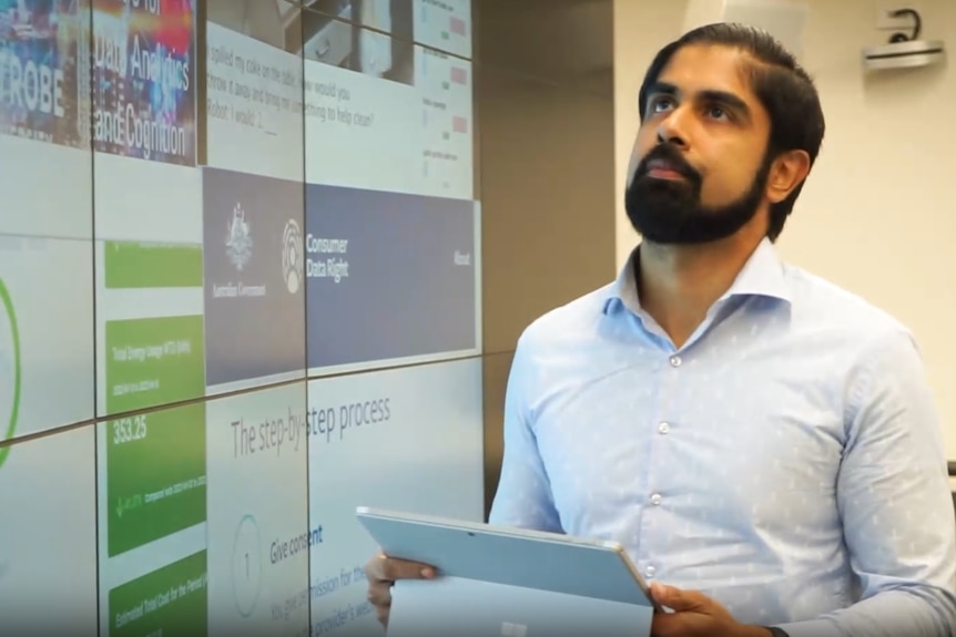 A man in a blue button-down shirt and a beard looks up holding a tablet device.