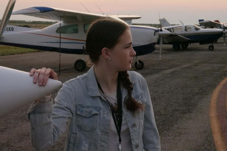 A young woman in a denim jacket stands with her hand on the nose of a Cessna light aircraft.