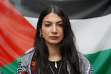 A woman with long brown hair stares directly in to a camera with a Palestinian flag behind her.