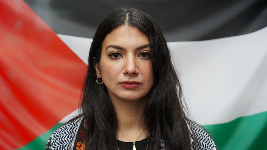 A woman with long brown hair stares directly in to a camera with a Palestinian flag behind her.