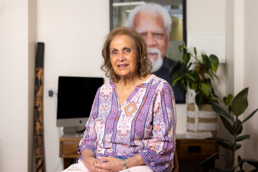 A black square with a circle in the centre with aunty naomi smiling. She is wearing a white, organise and white shirt