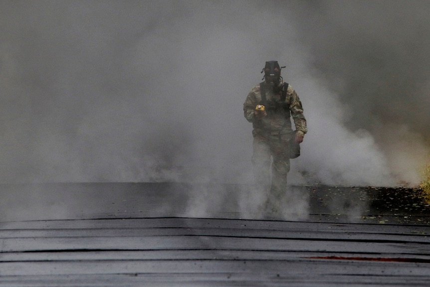 A man in a gas mask and dirty coveralls walks away from a crack in the road from which gas is streaming out