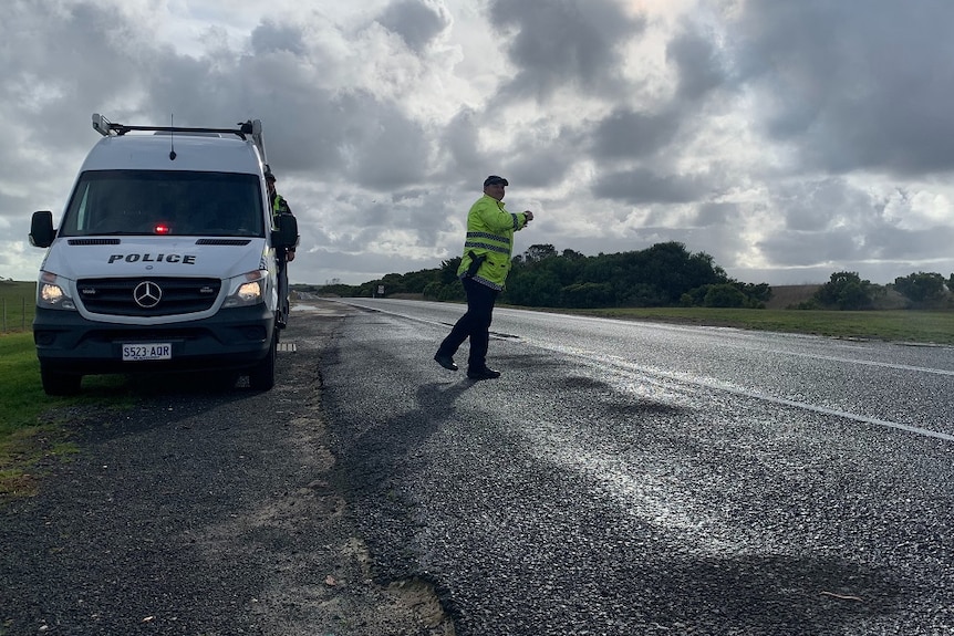 A police officer in hi-vis vest walks across a road with a parked police van behind him