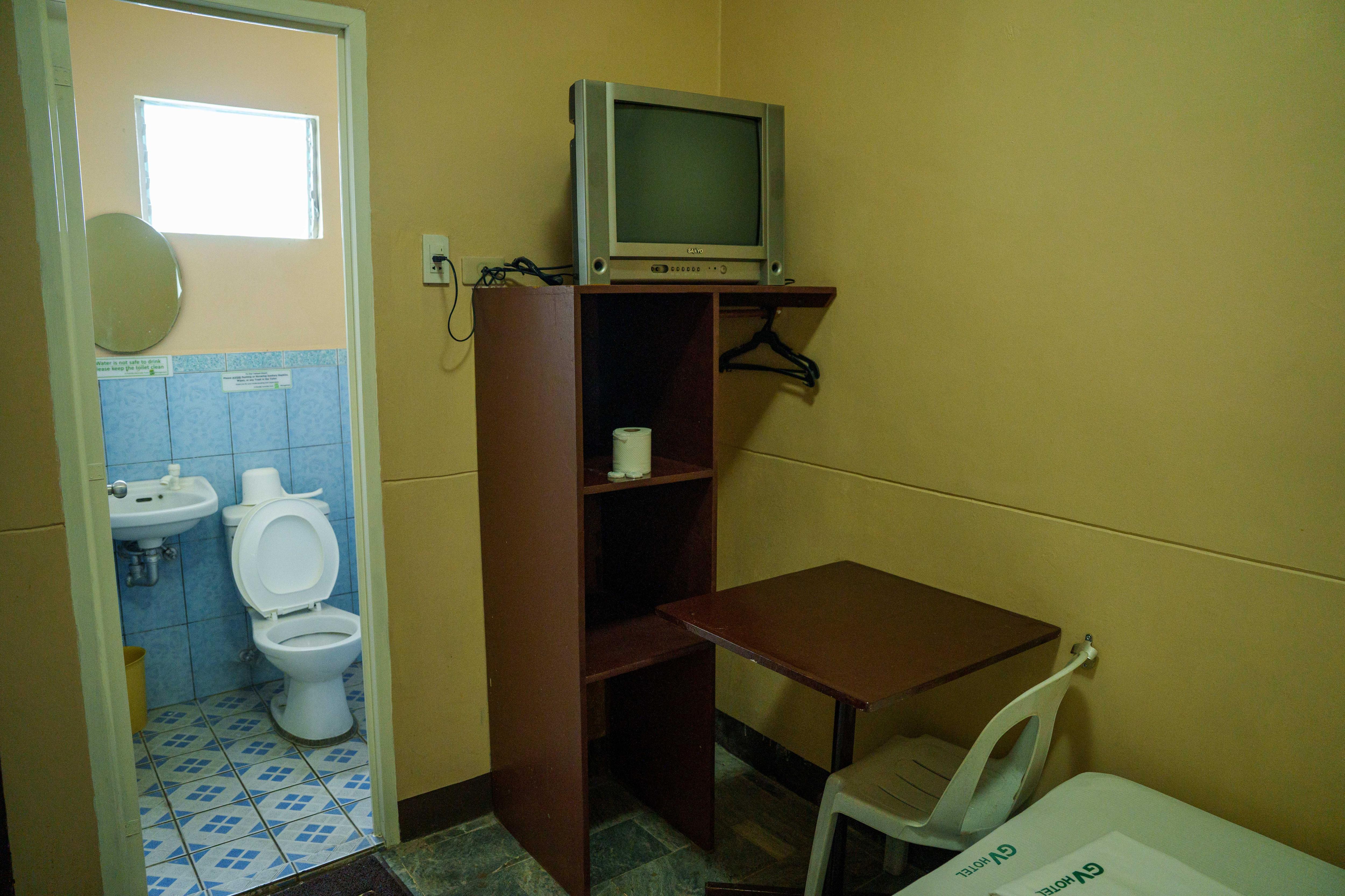O interior de um quarto de hotel sombrio, com uma pequena escrivaninha e cadeira de plástico, uma TV sobre uma mesa e um pequeno banheiro.