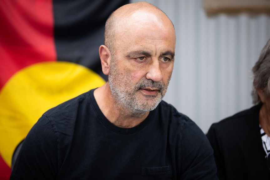 Man speaking in front of an Aboriginal flag.