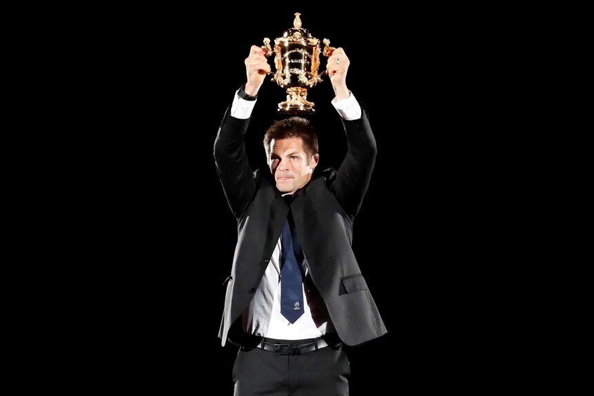 A male rugby union player wearing a suit, shirt and tie holds up a gold cup above his head.