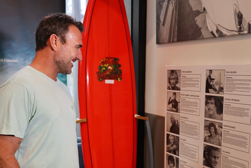 A man standing next to a surf board looking at pictures on a wall.