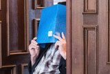 An elderly man in a wheelchair hides his face with a large blue booklet.