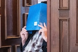 An elderly man in a wheelchair hides his face with a large blue booklet.