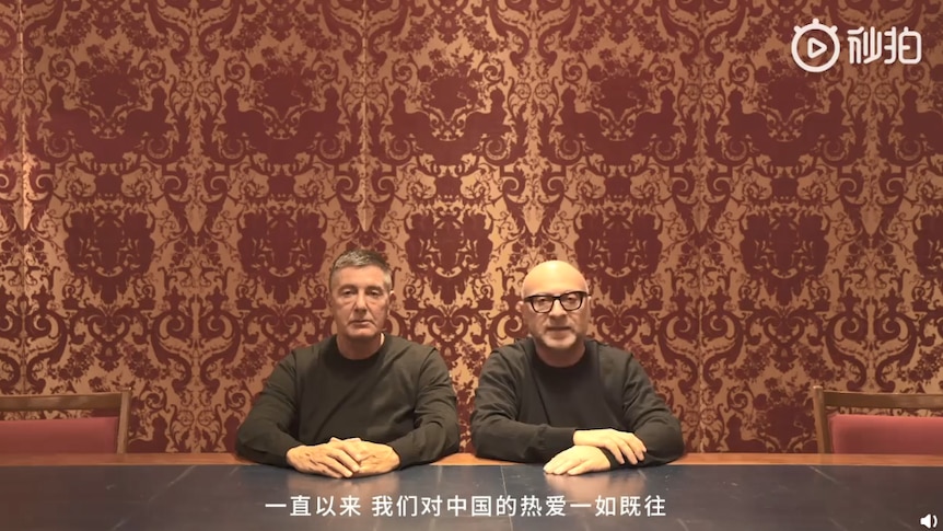 In front of an ornate red & gold wallpaper, Dolce & Gabbana founders clad in black sit behind black desk.