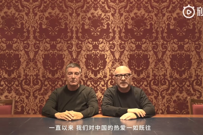 In front of an ornate red & gold wallpaper, Dolce & Gabbana founders clad in black sit behind black desk.