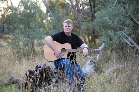 David Cox sitting in bushland on a large dead tree log with a guitar on his knee.