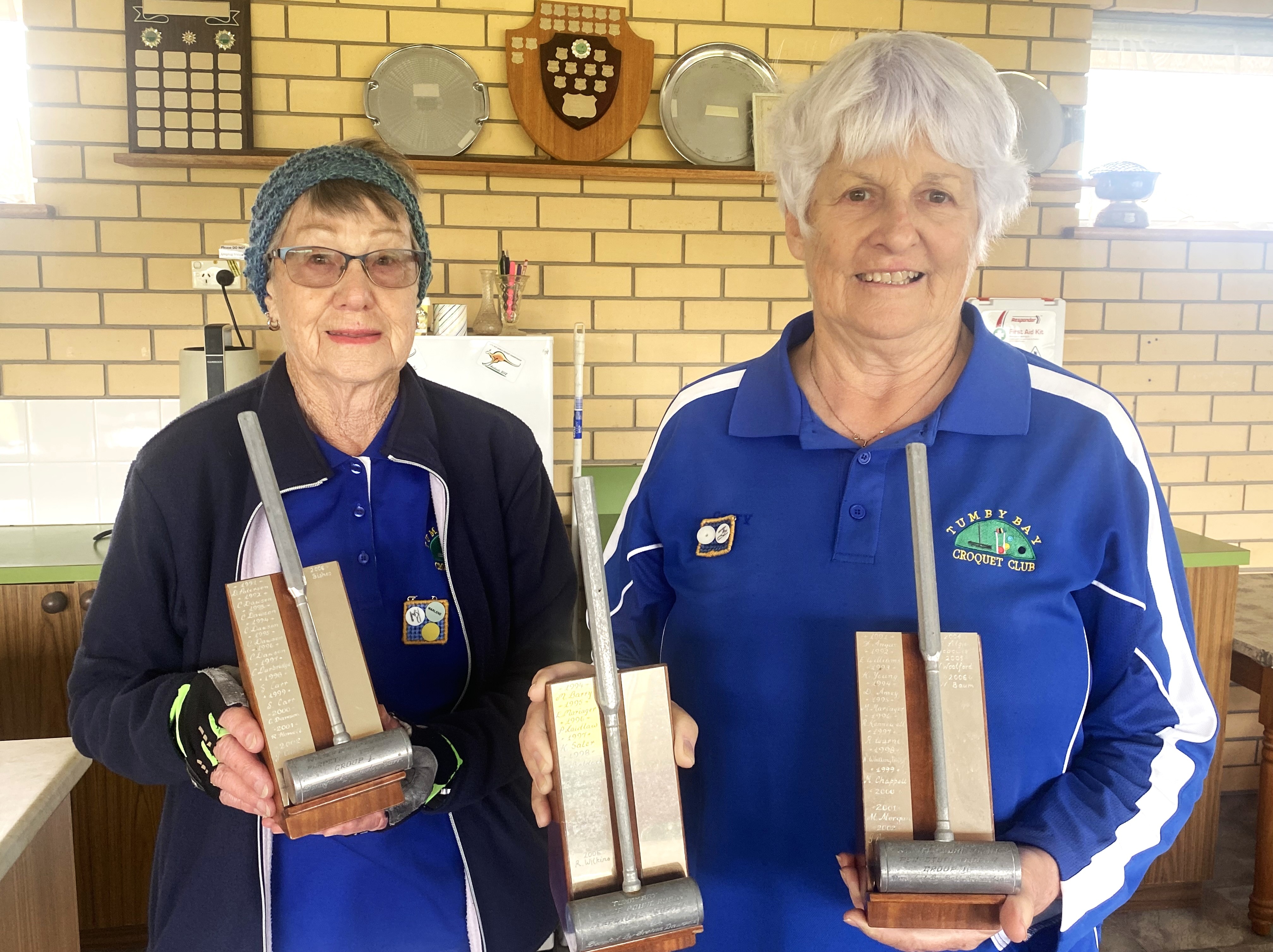 Duas mulheres segurando troféus, em frente a uma parede com uma prateleira de escudos
