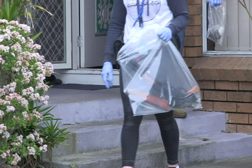 A military assault rifle in a plastic evidence bag is carried from a Melbourne home by a police officer.