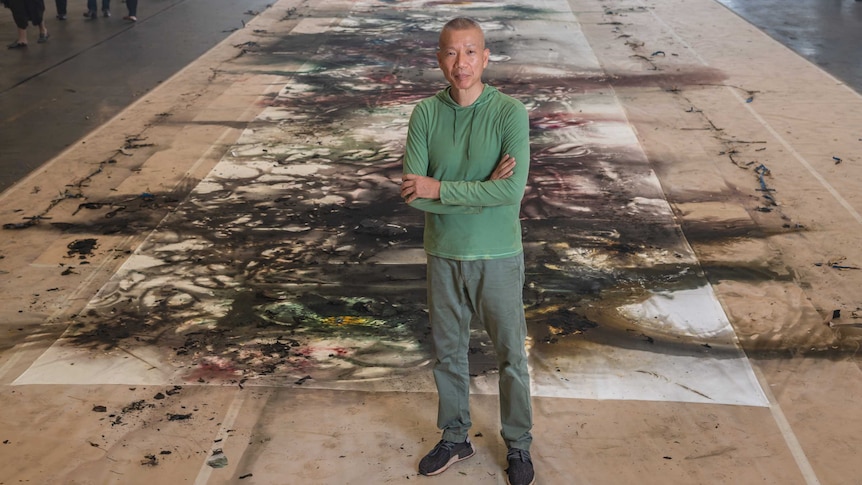 The artist stands arms folded in front of a silk canvas the length of the large room, covered in exploded gunpowder marks.