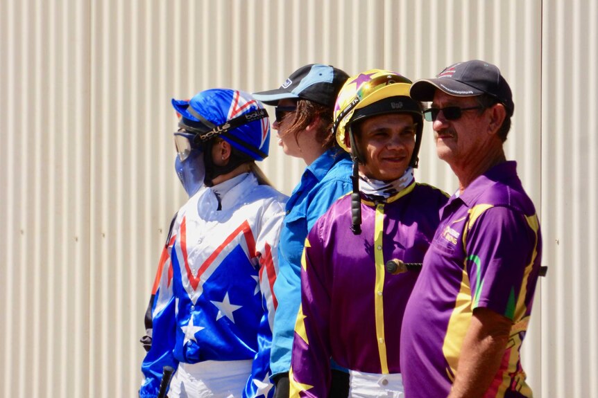 Two jockeys in silks and two trainers stand outside the jockeys rooms.