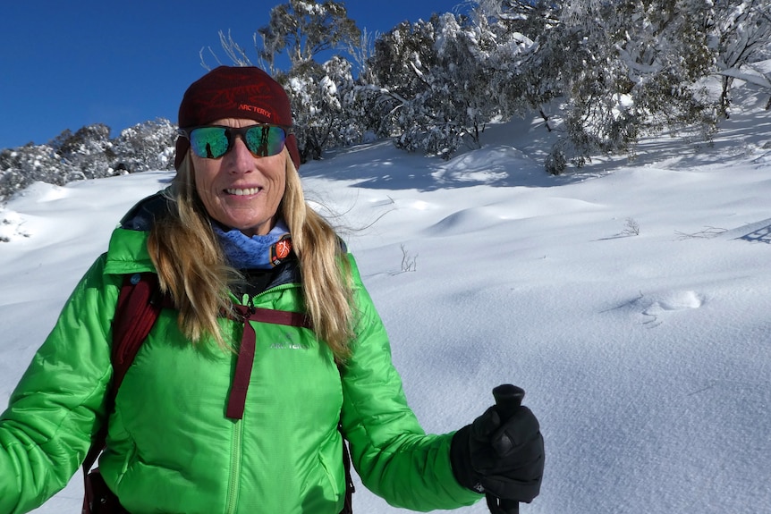 A woman wearing a green jacket and glasses stands in the snow.