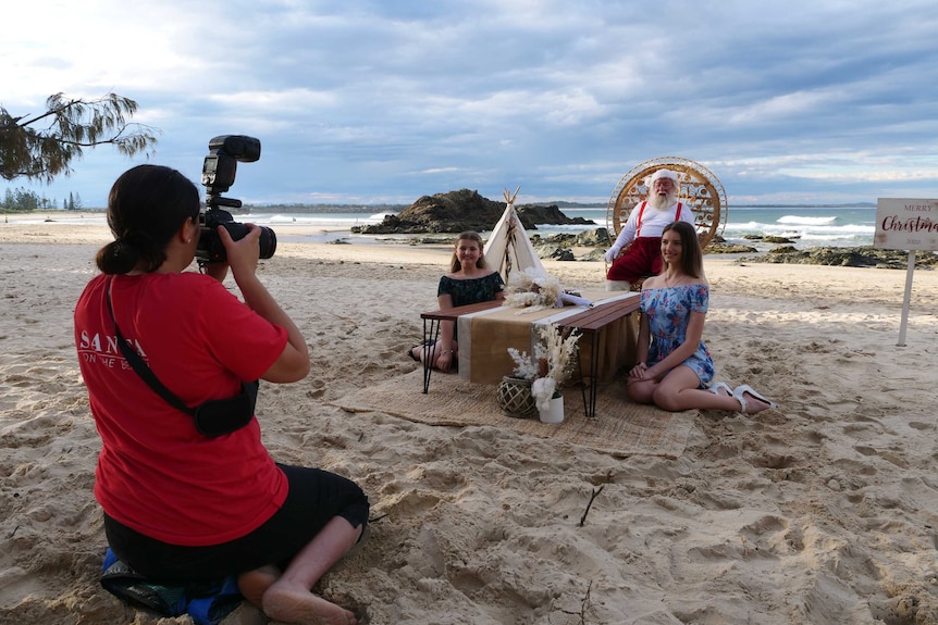 A photographer kneels on the sand taking a photo of two girls sitting on a beach, with Santa sitting on a chair behind them.