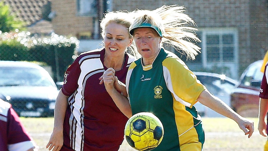 Two women run next to each other looking at a bouncing soccer ball that is in front of them at waist height.