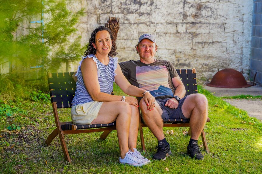 A couple in their 40s sit on chairs in a garden and smile at the camera with a stone wall behind them