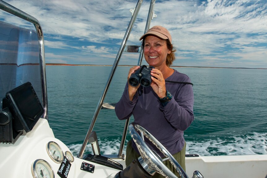 Dr Janet Mann on a boat on the ocean, smiling and holding a pair of binoculars in front of her