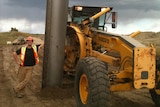 A smiling man in high-vis leans against a huge girder sticking out of the earth near a digger.