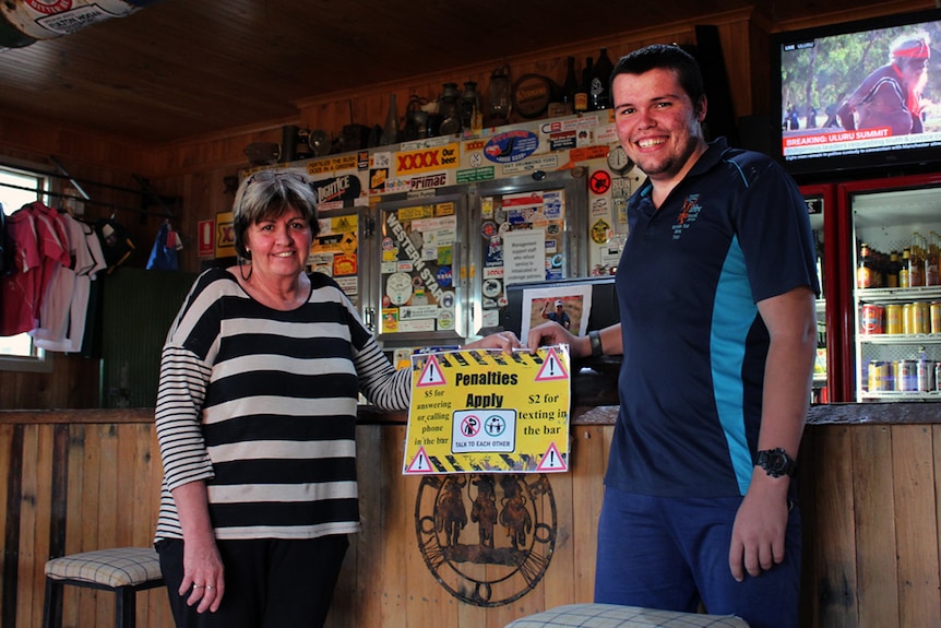 Marilyn Simpson and Ben Cross standing at the bar holding a sign that stipulates the charges for phone usage at the bar.