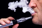 A man exhales a white substance while holding an e-cigarette device in front of his mouth.