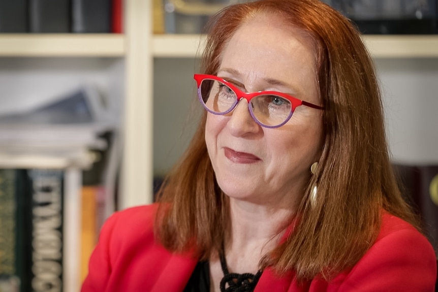 Woman with red hair in red blazer and red and purple glasses looks to left of frame.