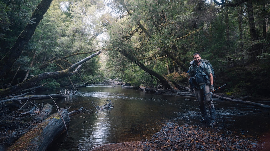 Man stands on bank of a river, smiling. In the background is wet eucalypt forest.
