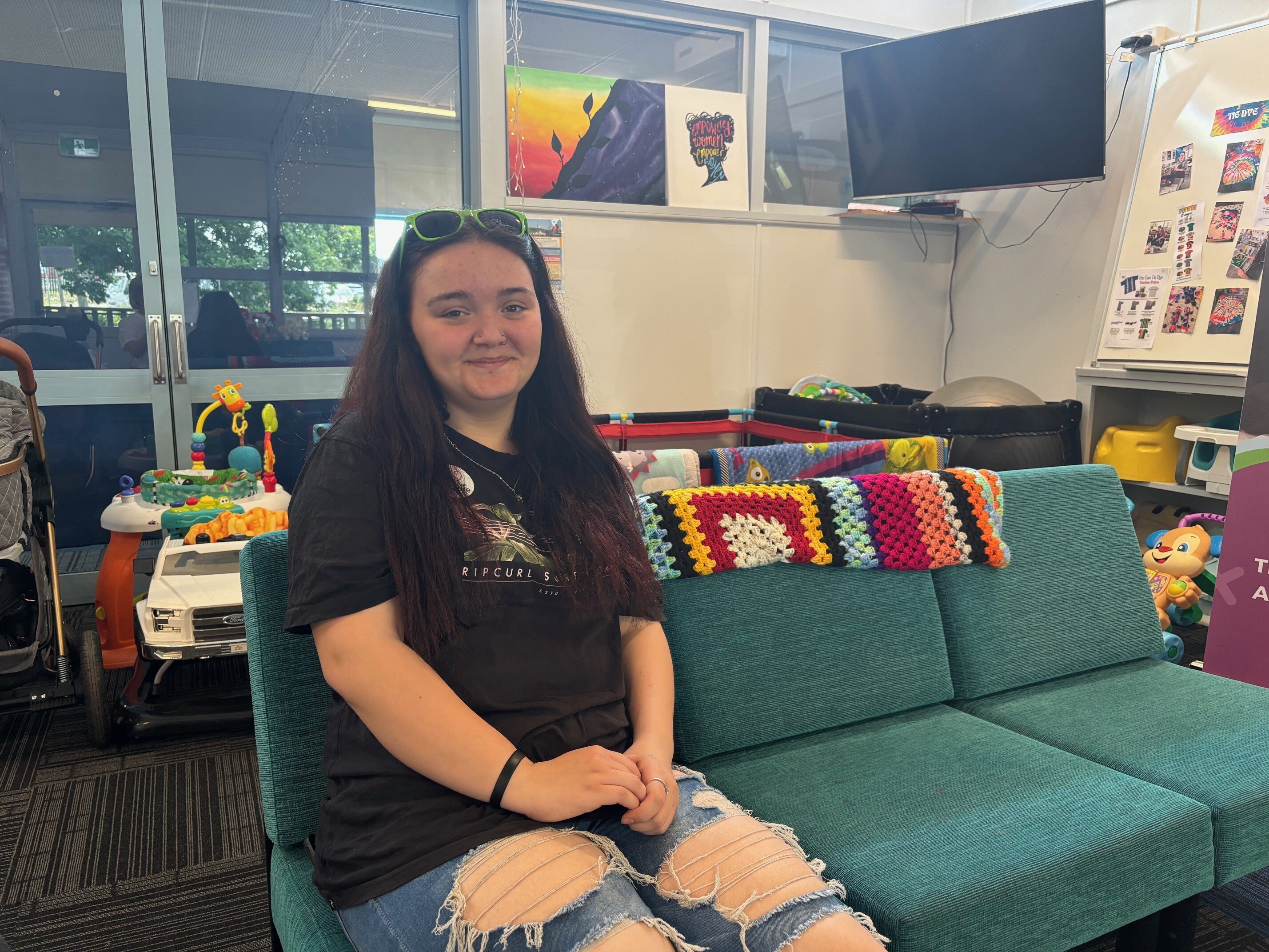 A smiling young girl, sunnies on top, black tee, torn jeans, green sofa, croche throw, children's toys at the back. 