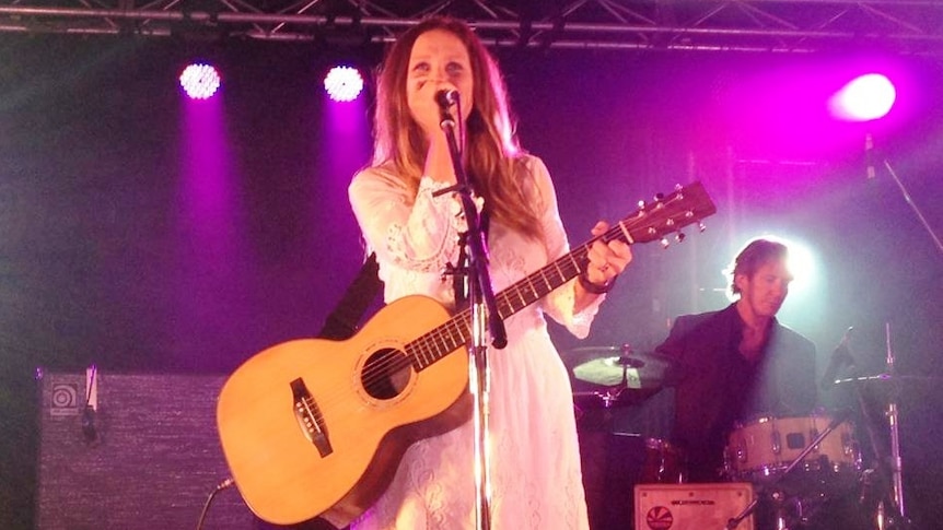 Kasey Chambers stands on stage with microphone in hand, pinkish lighting, she's smiling