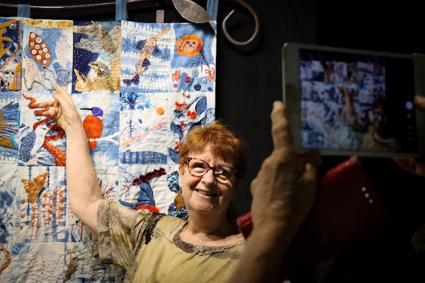 A lady points at her quilted piece and poses for a photo.