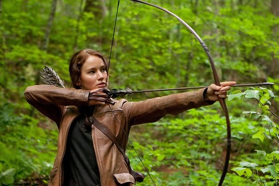 A woman wearing a brown leather jacket draws a bow and arrow.