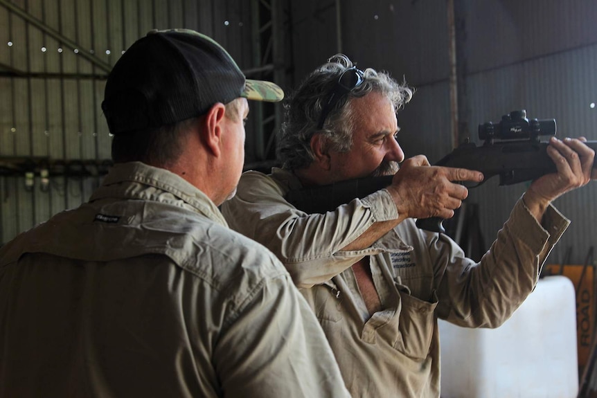A photo of Roger Matthews aiming a large rifle into the distance with a man watches on.