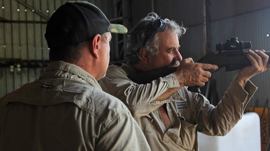 A photo of Roger Matthews aiming a large rifle into the distance with a man watches on.