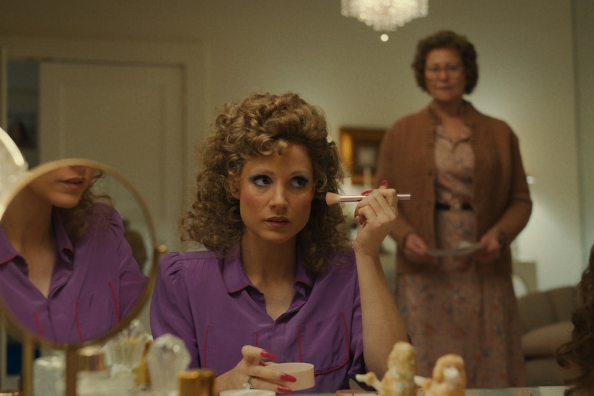 A blonde woman with an 80s perm sits applying make-up, ignoring an older woman standing behind her