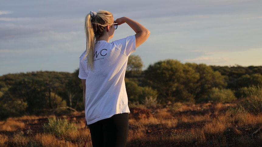 A blonde woman in a white t-shirt and tights looks over towards the sunset in the outback
