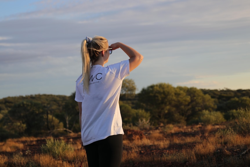 A blonde woman in a white t-shirt and tights looks over towards the sunset in the outback