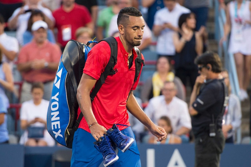 Nick Kyrgios walks off the court carrying his bag and shoes.