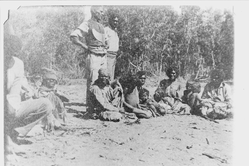 A black and white image of Aboriginal people sitting on the ground and standing circa 1913.