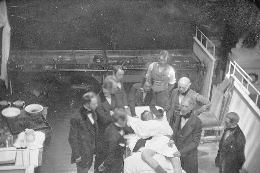 A group of men stand around a patient on a bed in a black and white recreation of the first operation under anaesthesia, 1846