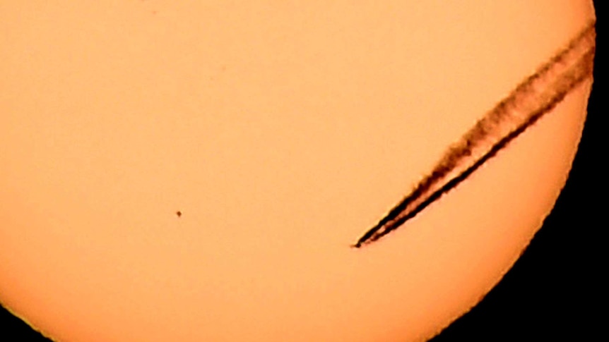 The planet Mercury appears as a black dot against the Sun as a plane, also silhouetted against the Sun, leaves a vapour trail.