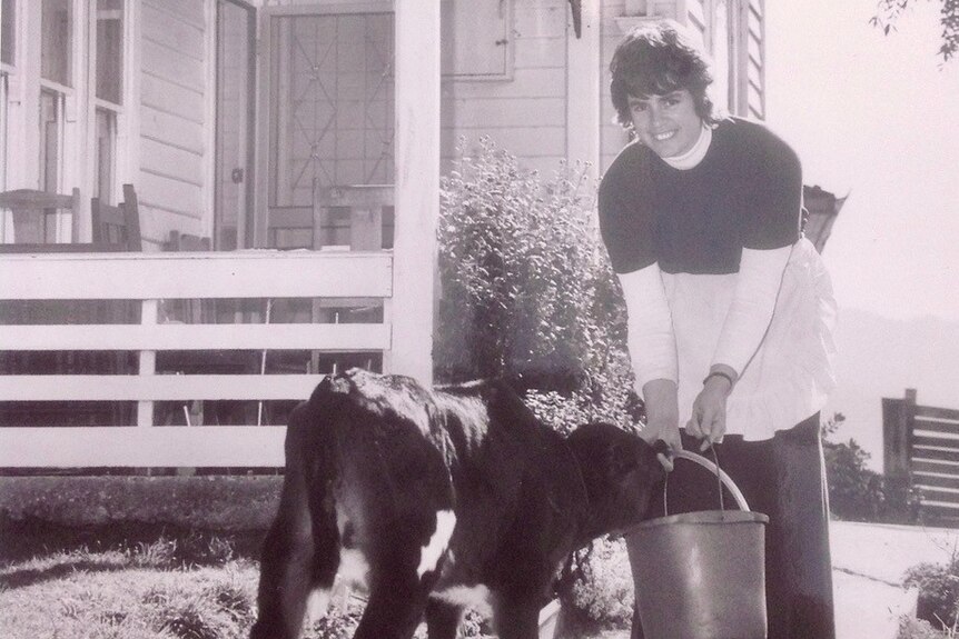 A black and white photo of a woman feeding a calf in front of a house.