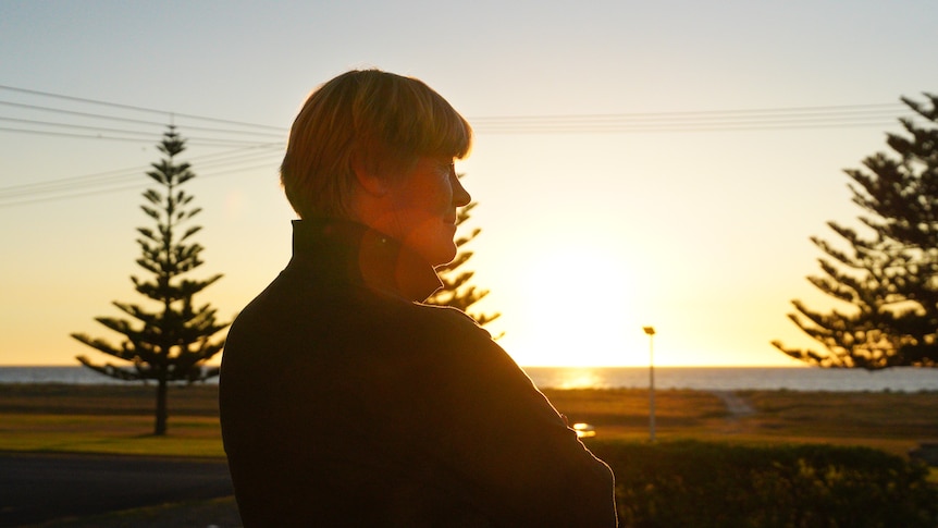 A woman stands on the foreshore of a beach lined with pine trees at sunset.