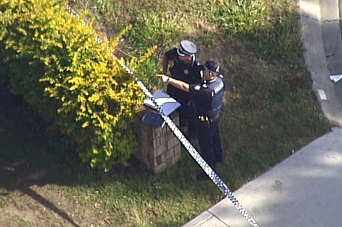 A police officer points past police tape while standing near the driveway of a house.