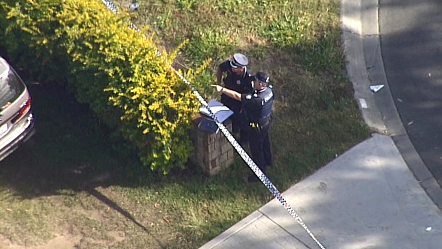 A police officer points past police tape while standing near the driveway of a house.