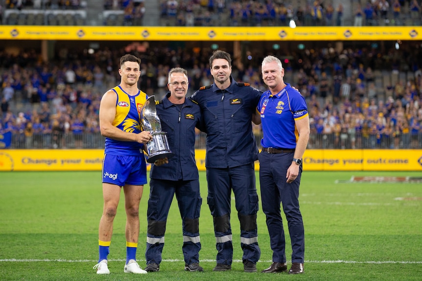 Liam Duggan and Adam Simpson of the West Coast Eagles standing with two emergency services workers holding a trophy.